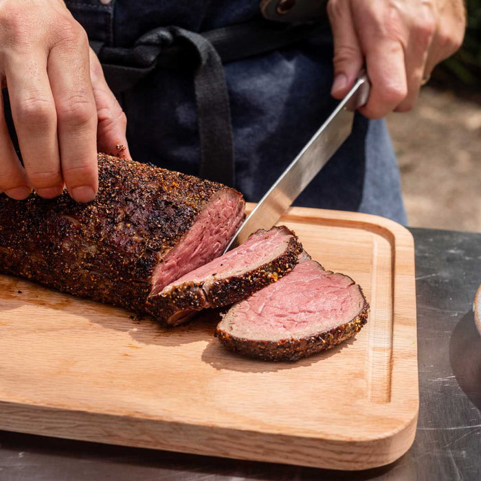 Una mano tiene la parte superiore di un filet mignon cucinato a cottura lenta, l'altra mano affetta la bistecca con un grosso coltello su un tagliere di legno sopra un Tavolo modulare Ooni.