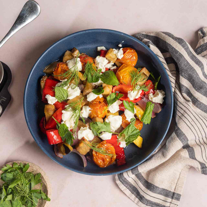 Zucca, melanzane, cipolle, pomodori ciliegini e peperoni dolci arrostiti a fuoco lento in una teglia per arrosti Ooni su un tavolo accanto a una ciotola che contiene verdure cucinati a fuoco lento con feta ed erbe fresche.