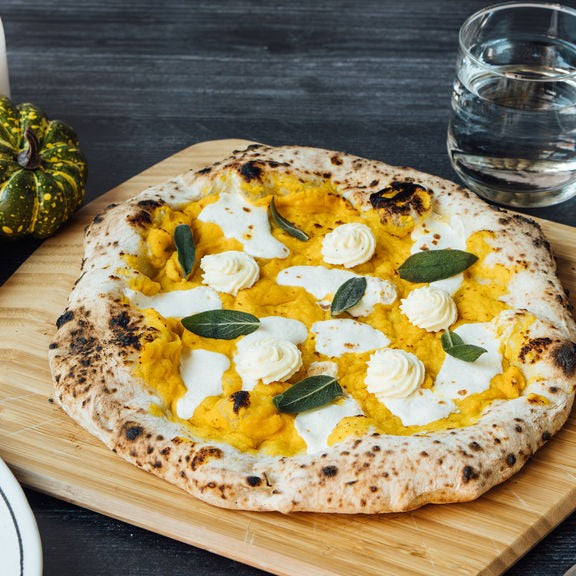 Pizza con zucca violina, ricotta e salvia fritta su un tagliere per pizza in bambù Ooni su un tavolo accanto a piatti, un bicchiere d'acqua e zucche decorative.