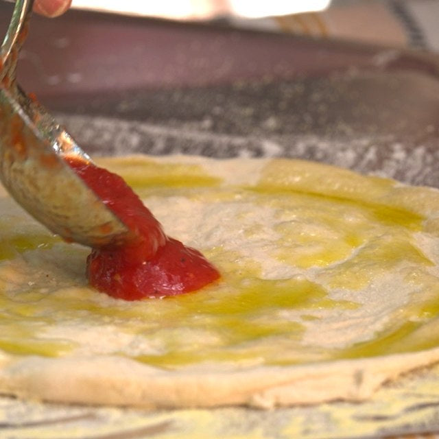 La salsa di pomodoro viene versata con un mestolo su un impasto per pizza condito con olio di oliva, steso su una pala per pizza Ooni.
