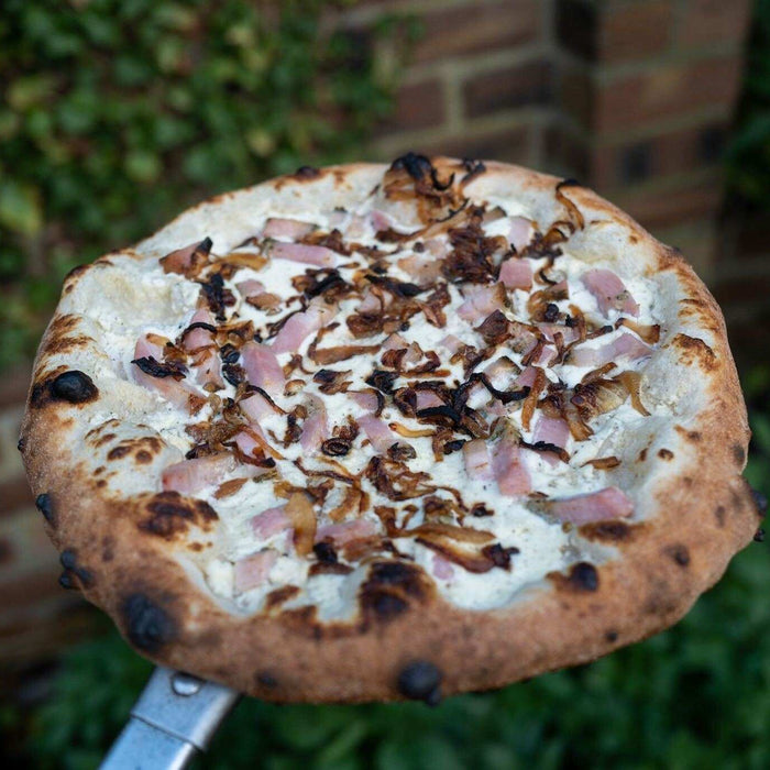 Pizza in stile napoletano ispirata alla tarte flambée alsaziana con crème fraîche, formaggio frais, cipolle, pancetta e scalogno su una pala per pizza Ooni.